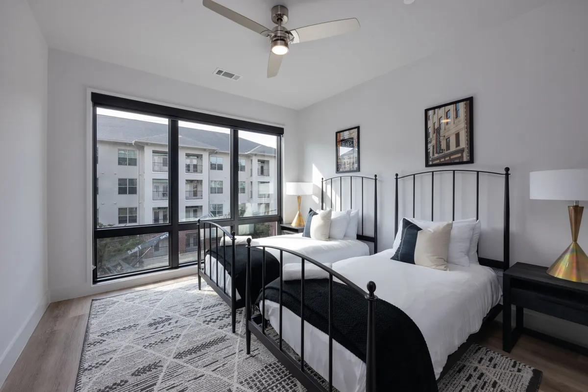 Featuring two twin beds with crisp linens, a large window letting in ample daylight, decorative wall art, and a nightstand with a lamp, this room radiates calm and flexibility—ideal for friends, family, or business guests.