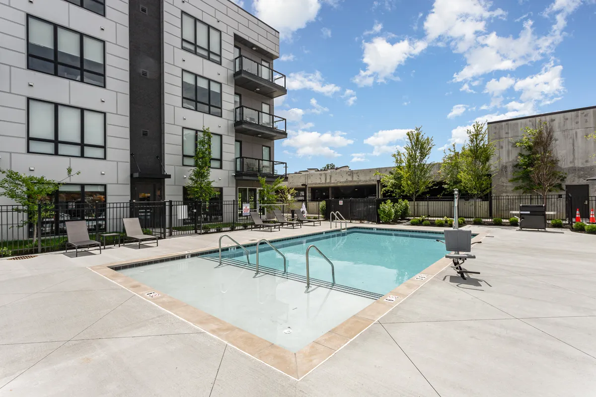 The image captures a modern outdoor swimming pool area surrounded by lounge chairs and landscaped greenery, with the apartment building in the background. The pool is inviting, with ample space for relaxing, sunbathing, or gathering with friends.