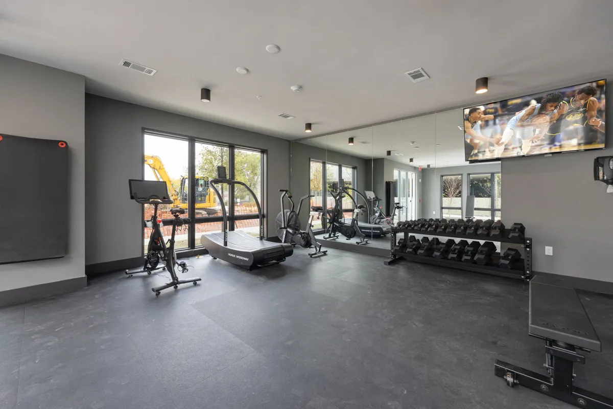 A bright, modern fitness center interior with high-end exercise equipment, large windows letting in natural light, mirrored walls, and a TV screen. The space conveys an energetic, focused mood and is designed for both solo workouts and group fitness classes, aligning with the state-of-the-art gym mentioned in the text.