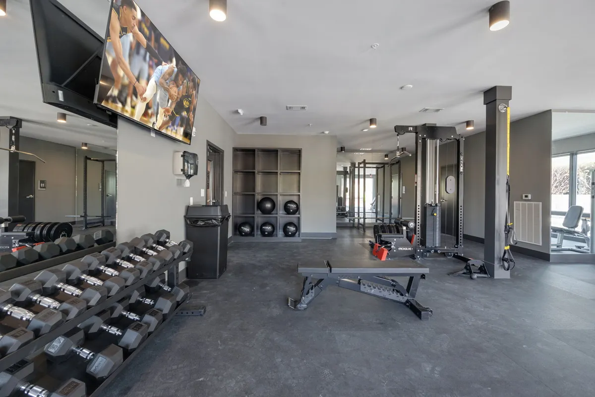 This interior shot features a fully equipped fitness center with a dumbbell rack, weight bench, cable machine, exercise balls, and a wall-mounted television. The space is bright and motivating, designed for comprehensive workouts.