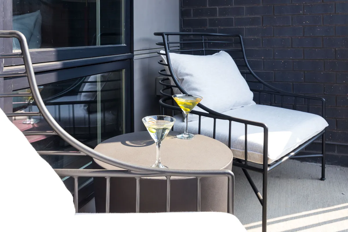 An exterior patio scene with two modern chairs and a small round table set between them, upon which rest two martini glasses. Surrounded by open sky and cityscape hints, the scene suggests relaxed outdoor socializing, sunset views, and a strong sense of urban leisure.