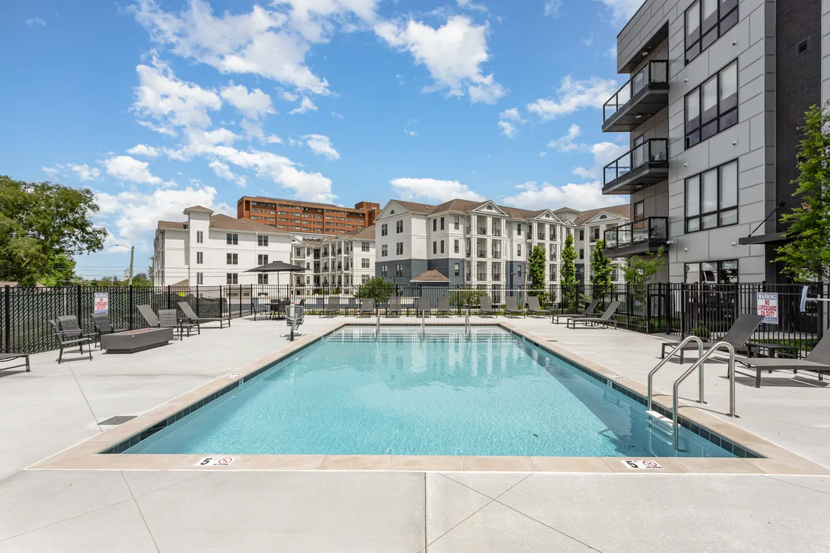 An exterior view of the swimming pool area, showing the pool itself, surrounding lounge area, and nearby residential buildings. People can be seen swimming, sunbathing, and socializing, evoking a leisurely, communal mood.