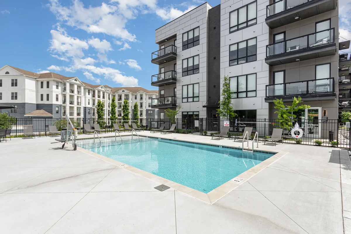 A vibrant exterior shot featuring a sparkling swimming pool surrounded by landscaped gardens and the balconies of modern apartment buildings. Lounge chairs are set up for sunbathing or relaxation, and the setting exudes a resort-like ambiance. The scene suggests opportunities for both social gatherings and tranquil solo moments, enhanced by sunset lighting.